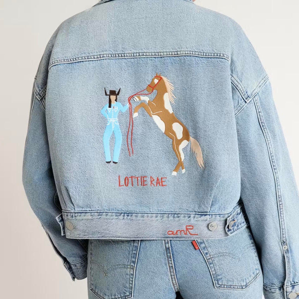 Custom chainstitch embroidery denim jacket created for Lottie Rae x Levi’s, featuring a cowgirl in a light blue western outfit beside a rearing brown and white horse, with LOTTIE RAE stitched in red on the back