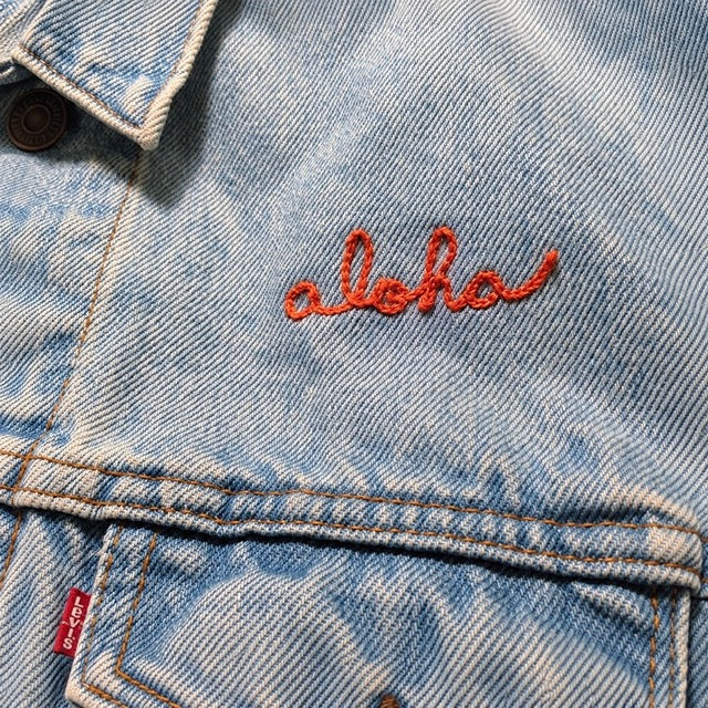Close-up of custom chainstitch name embroidery by amR on a vintage-wash Levi's denim jacket, featuring Aloha in vibrant orange cursive lettering with a delicate heart-shaped detail, stitched above the front chest pocket
