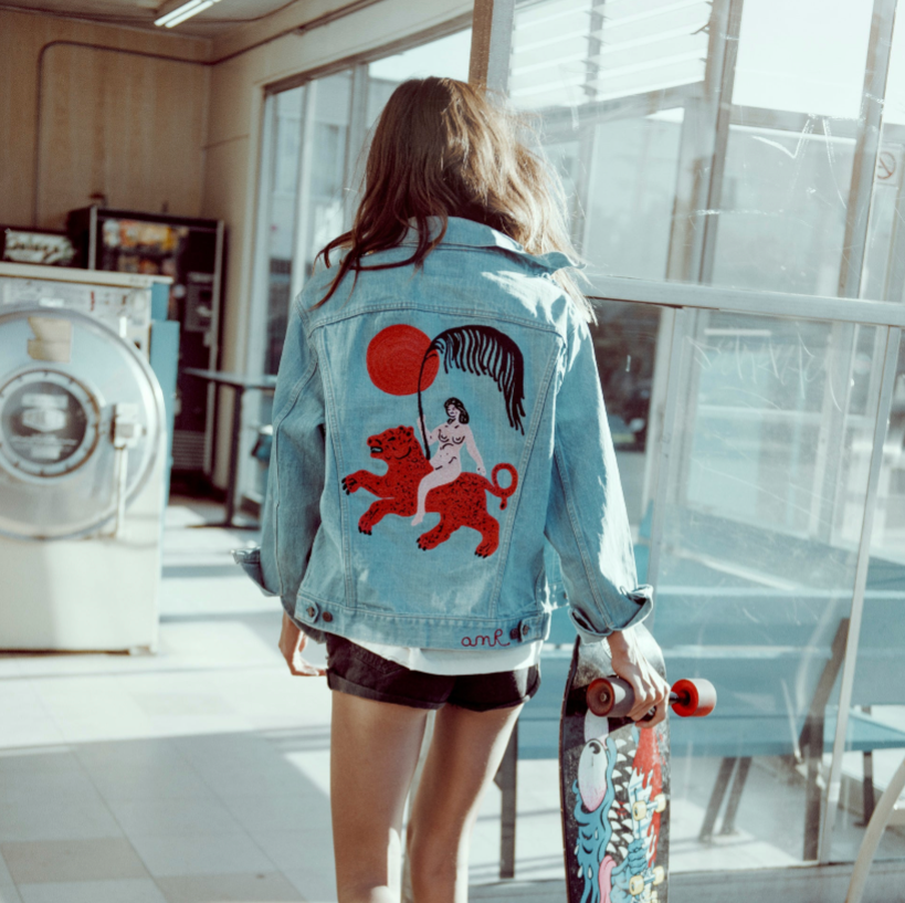 Custom chainstitch embroidered light blue denim jacket by amR, featuring a bold illustrated back design with a red tiger, a seated figure, a crimson sun and a curved black-and-white motif, worn by a woman holding a skateboard.