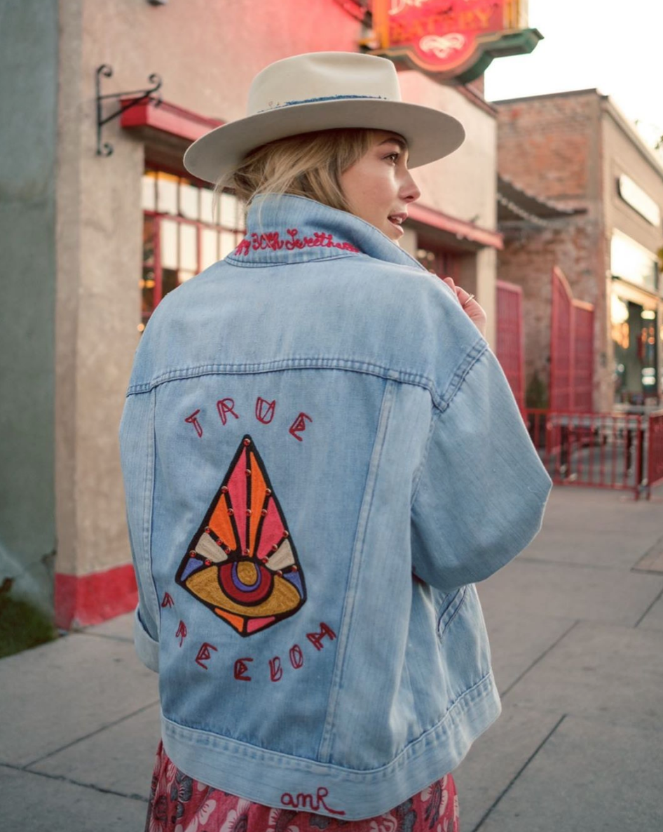 Custom chainstitch embroidered light blue denim jacket worn with a western hat, featuring a colorful geometric eye motif, red TRUE FREEDOM lettering, intricate stitched details and custom collar embroidery by amR