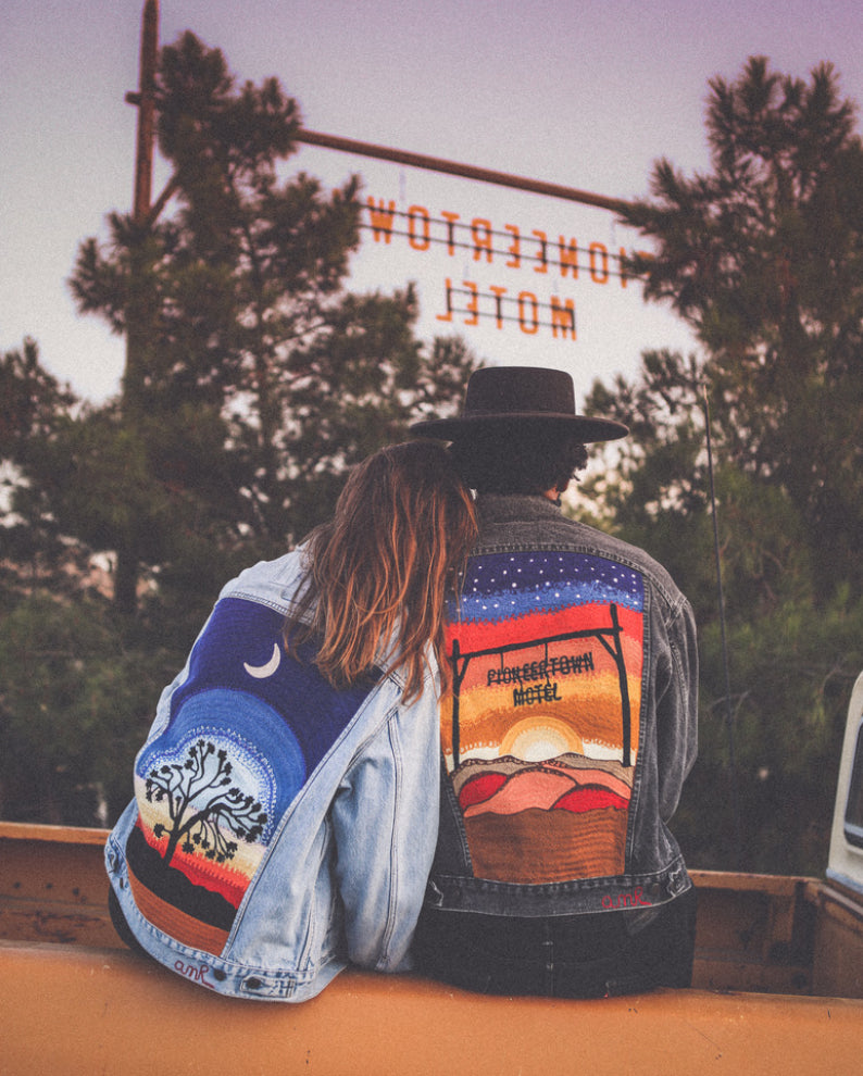 Two custom chainstitch embroidered denim jackets by AMR, featuring colorful Joshua Tree desert landscapes, a crescent moon, starry skies and a Pioneertown Motel sunset design, worn outdoors beneath the motel sign