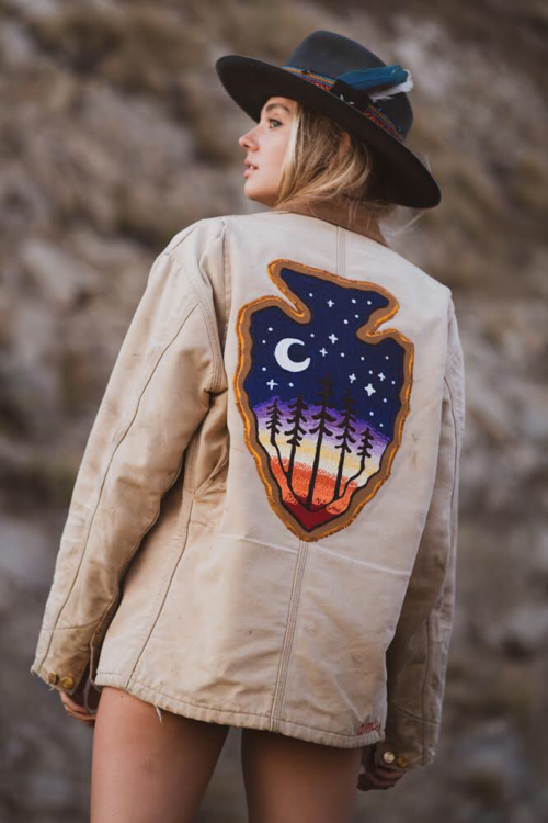 Lifestyle photo of a woman wearing a custom embroidered beige jacket featuring a large moonlit forest back design with a crescent moon, stars, pine trees and a sunset gradient, shown outdoors with a black hat