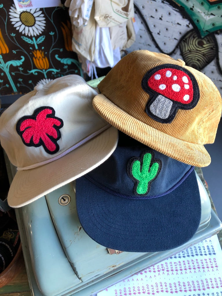 Custom embroidered patches by amR on three vintage-style caps, featuring a red and white mushroom on mustard corduroy, a bright green cactus on navy blue and a pink flower on cream canvas, with raised textured stitching and bold black borders