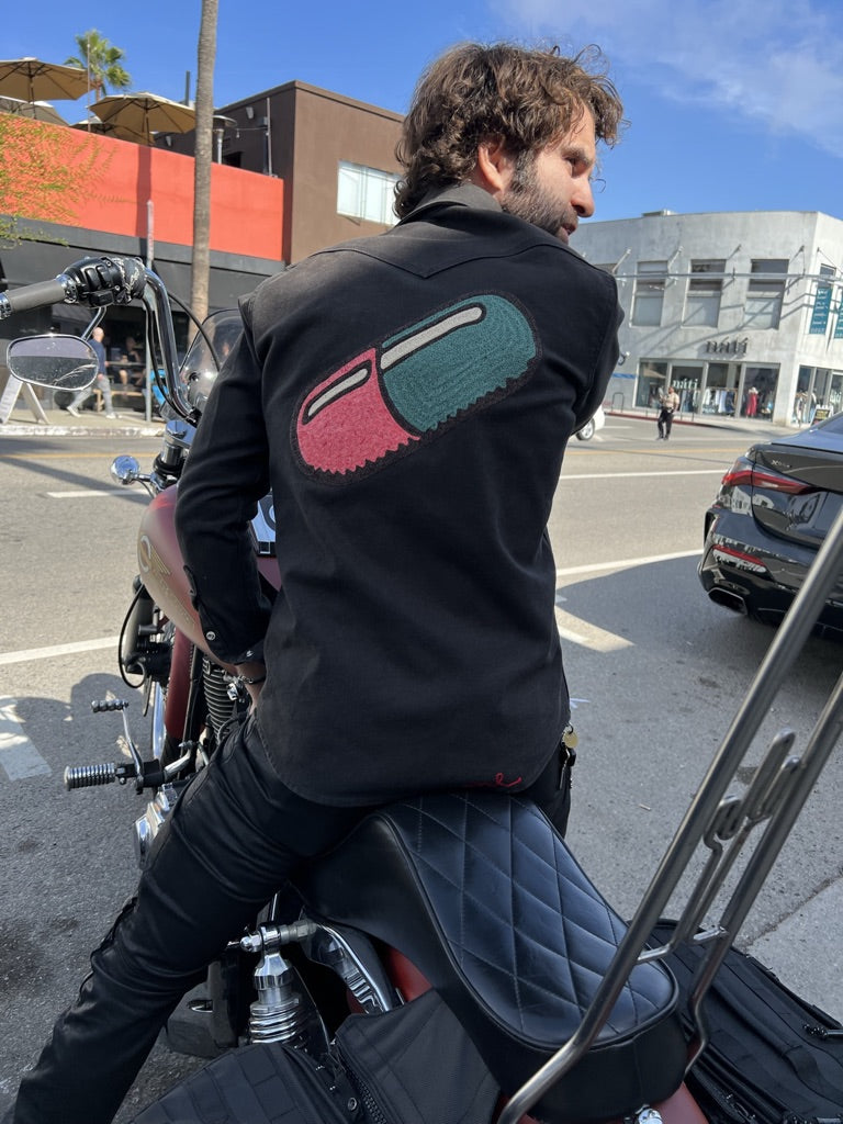 Custom chainstitch embroidery on a black denim western shirt worn by a motorcycle rider, featuring a large pink and teal capsule back design with textured stitching and bold black outlines, photographed on a city street by amR in Los Angeles