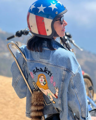 Woman wearing a custom chainstitch embroidered denim jacket by amR in Los Angeles, with illustrated back artwork and a motorcycle helmet