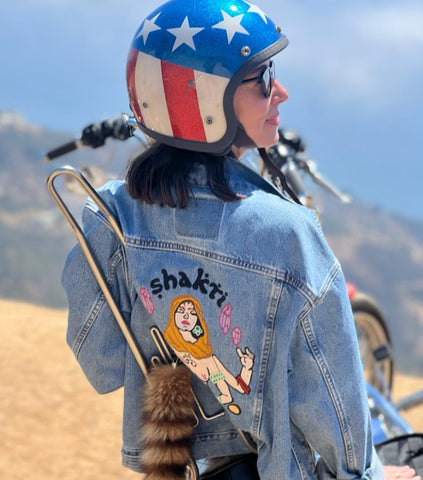 Woman wearing a custom chainstitch embroidered denim jacket by amR in Los Angeles, with illustrated back artwork and a motorcycle helmet