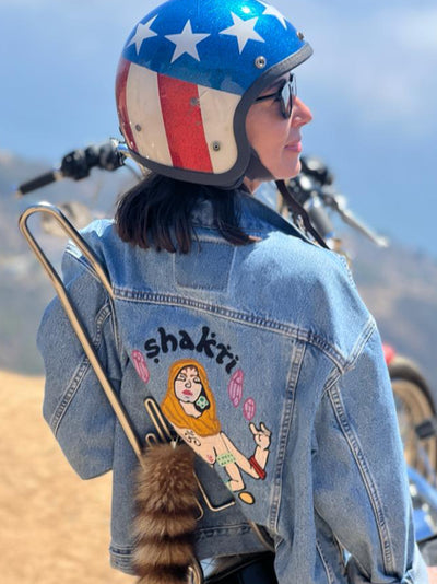 Woman wearing a custom chainstitch embroidered denim jacket by amR in Los Angeles, with illustrated back artwork and a motorcycle helmet