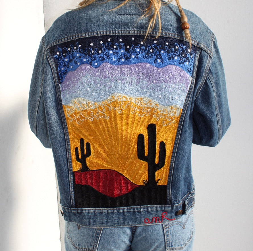 Custom chainstitch embroidery on a vintage-style light blue denim jacket, featuring a full-panel Southwestern desert landscape with black saguaro cacti, radiant golden sunbeams, red desert hills, layered lavender and pale blue mountains, and a star-filled midnight blue sky, crafted by amR