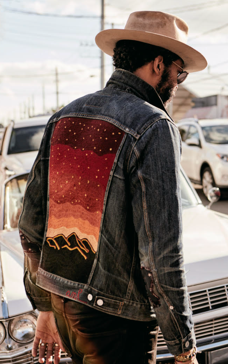 Custom chainstitch embroidered dark denim jacket by AMR, featuring a large desert landscape back design with a star-filled red sky, warm sunset gradients and black mountain silhouettes, worn outdoors with a western hat