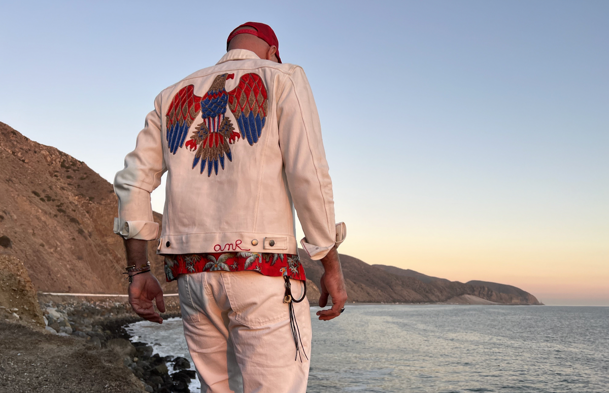 A man viewed from behind wearing a cream-colored denim jacket with a large embroidered eagle design in red, blue, and gold on the back, standing on a rocky coastline at sunset.