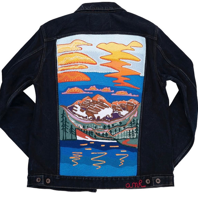 Custom chainstitch embroidery on a dark indigo denim jacket, featuring a colorful mountain sunset landscape with snow-capped peaks, pine forests, a blue lake and intricate stitched details by amR