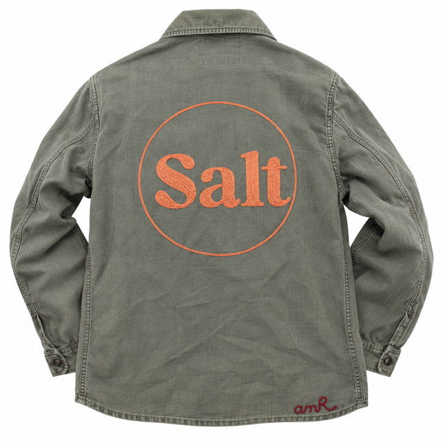 Custom chainstitch embroidery on a faded olive green denim shirt, featuring oversized orange Salt lettering with textured fill stitching inside a circular embroidered outline, finished with a red AMR signature near the hem