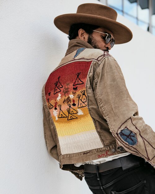 Western-inspired custom chainstitch embroidery on a tan workwear jacket, featuring a large desert sunset back design with geometric teepee motifs, scattered desert plants, warm ombre stitching and embroidered sleeve details, worn with a wide-brim hat