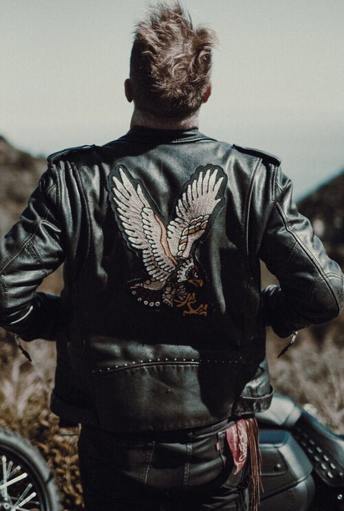 Custom leather jacket embroidery by AMR, featuring a large eagle back design with raised ivory feathers, black outlines and golden talons on a vintage black biker jacket worn by a motorcyclist outdoors beside a motorcycle