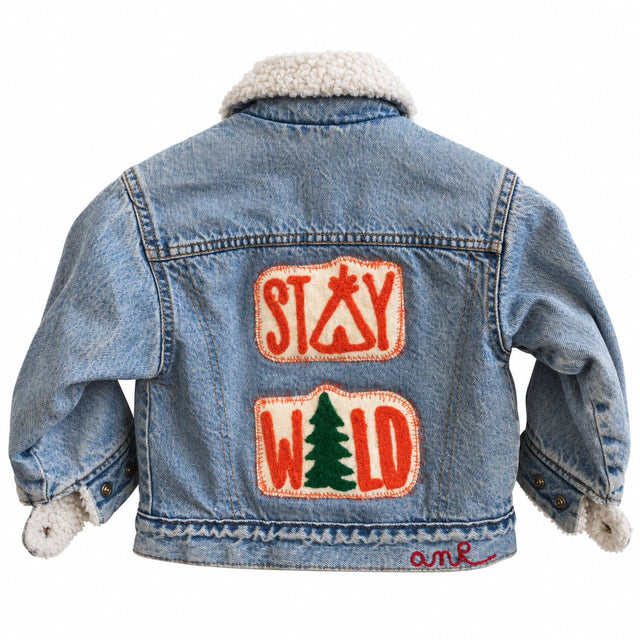 Bespoke chainstitch embroidery on a light blue kids' denim jacket with a cream sherpa collar, featuring playful STAY WILD lettering in orange on two textured back patches, a green pine tree motif and contrasting stitched borders, custom made by amR