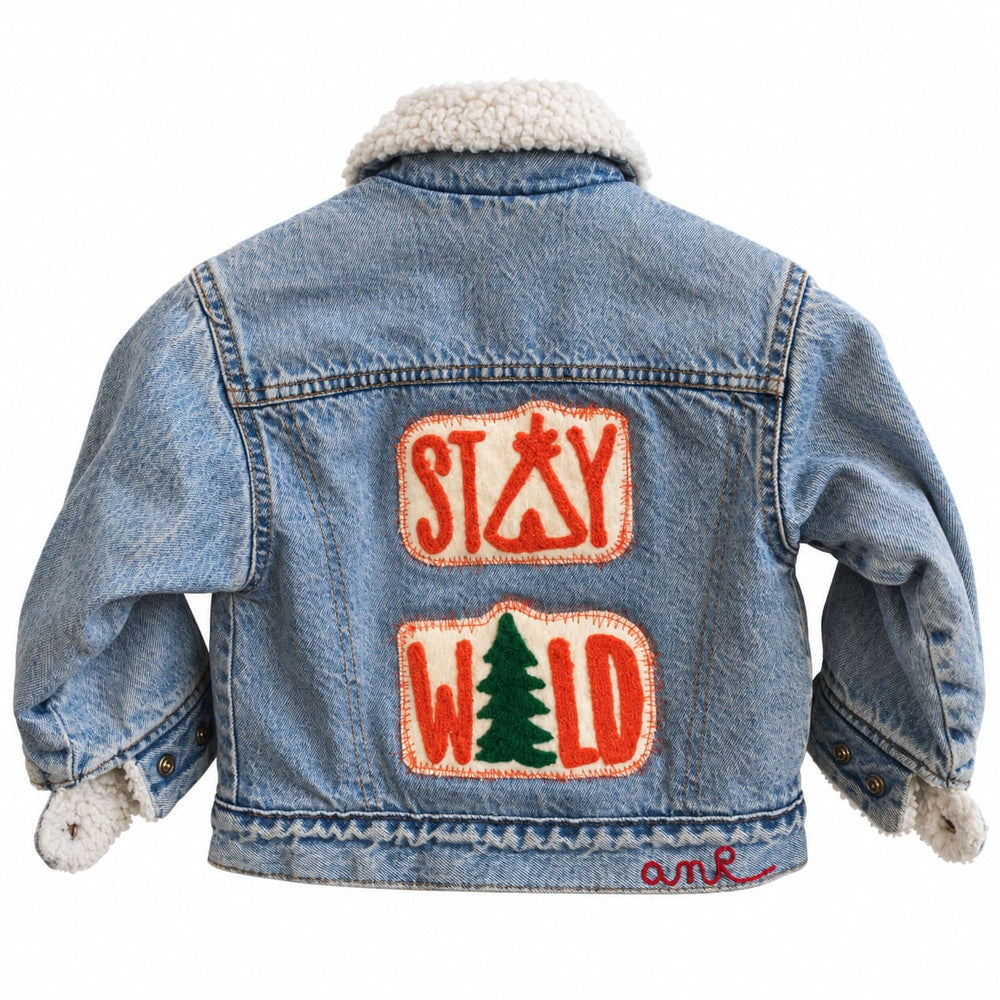 Bespoke chainstitch embroidery on a light blue kids' denim jacket with a cream sherpa collar, featuring playful STAY WILD lettering in orange on two textured back patches, a green pine tree motif and contrasting stitched borders, custom made by amR