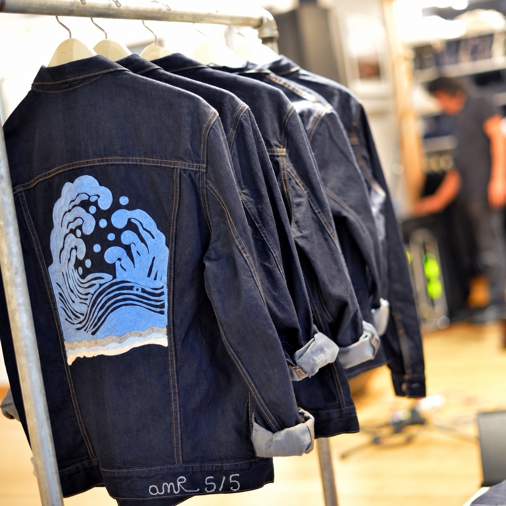 Limited-edition Ron Herman x amR denim jacket collection displayed on a retail clothing rack, featuring bespoke chainstitch embroidery with a large Japanese-inspired ocean wave design in shades of blue, cream and sand on dark indigo denim, with numbered amR 5/5 detailing